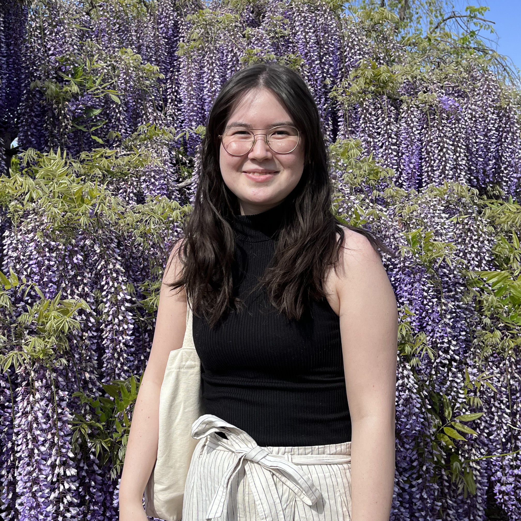 A bright, outdoor photo of Kyoko Telfer. She is standing in front of a forest of trees with cascading purple flowers. She has big glasses with thin rims and long, dark hair.