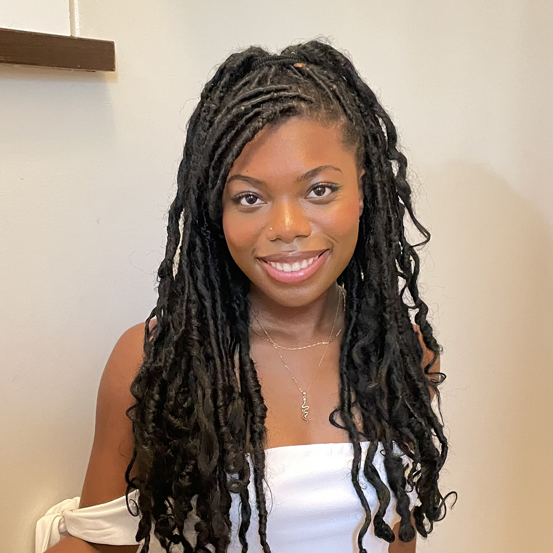 A smiling headshot of CURL Fellowship winner Lewa Babalola. She has long, dark hair tied up in pretty, curly twists. She wears a white off-shoulder top.
